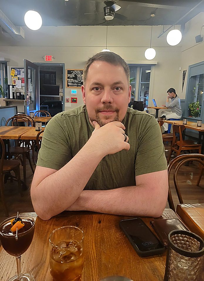 Gregg Roman at a cafe table at night, arms folded, chin on fist, looking at the camera