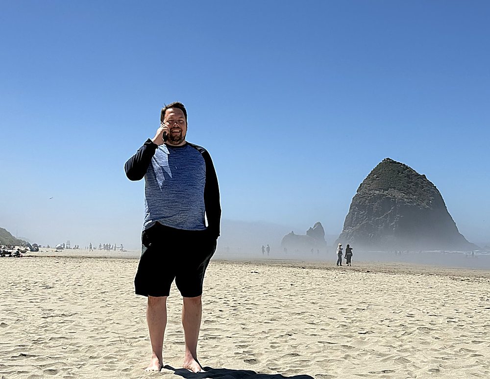 Gregg Roman barefoot on the sand at Cannon Beach, Oregon, taking a phone call with Haystack Rock in the mist behind him