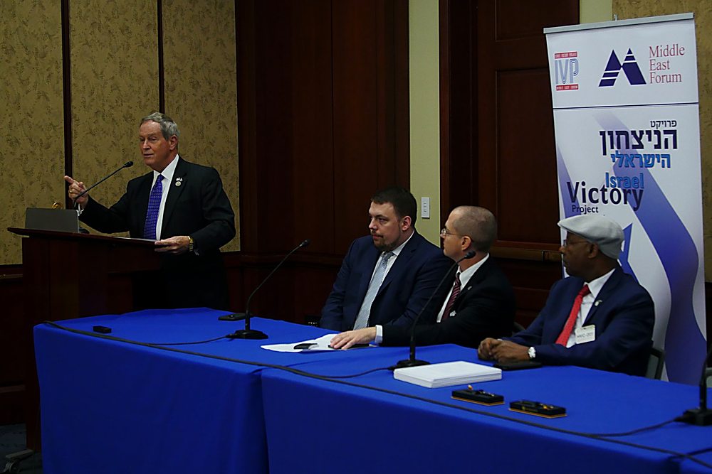 A congressman speaks at a podium while Gregg Roman sits on the panel at an Israel Victory Project briefing on Capitol Hill
