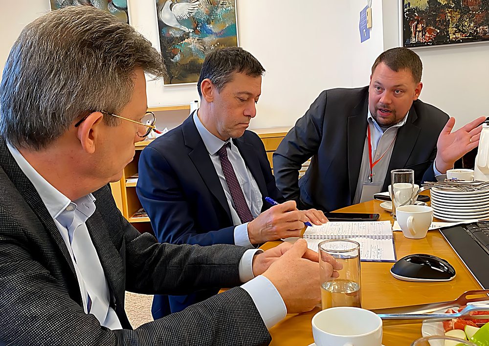 Gregg Roman gestures mid-explanation at a working meeting around a table with two colleagues taking notes