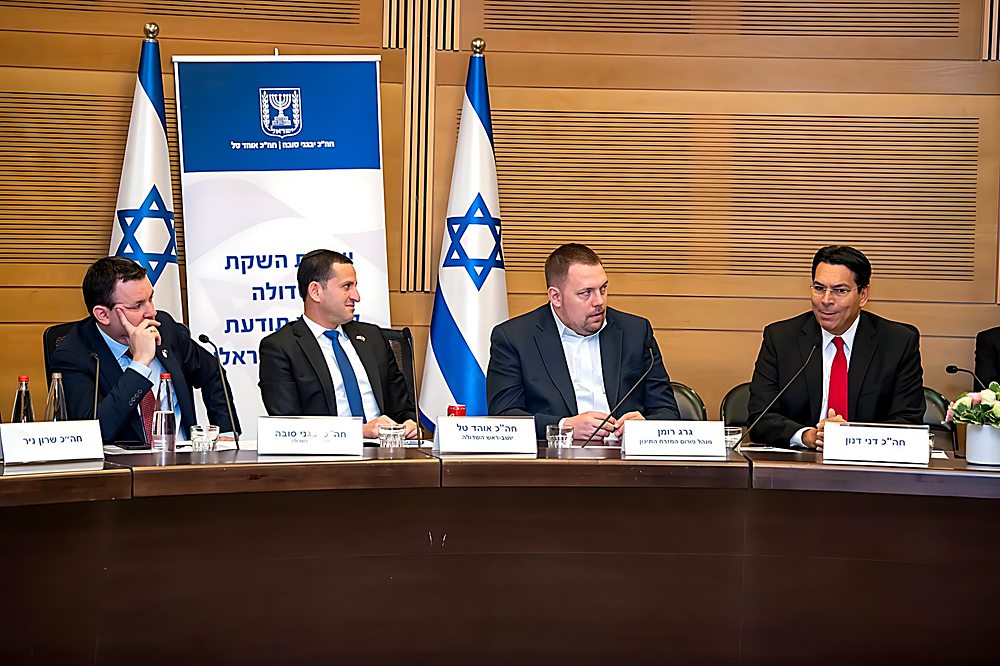 Gregg Roman seated behind his nameplate at a Knesset caucus table, beside MK Danny Danon
