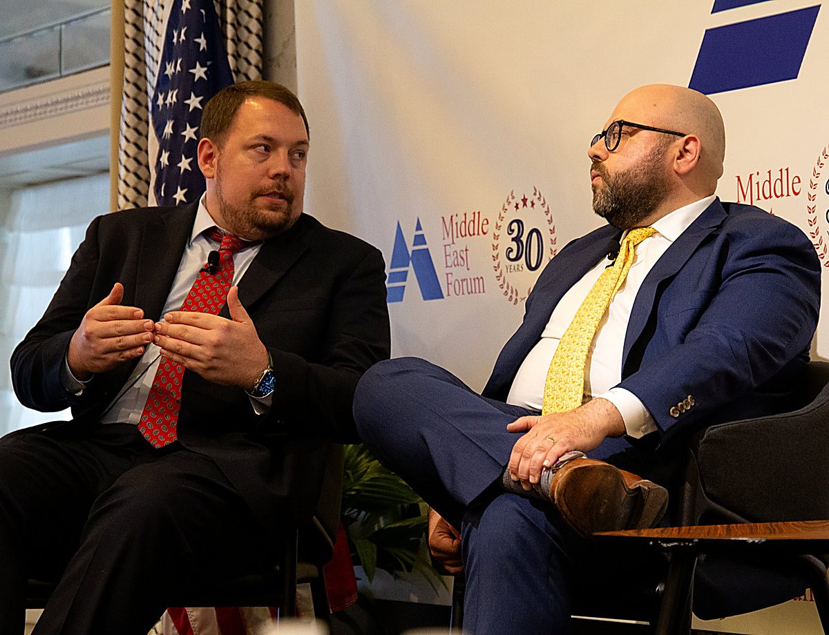 Gregg Roman gestures while making a point on a Middle East Forum panel, seated beside a fellow panelist with a U.S. flag behind them