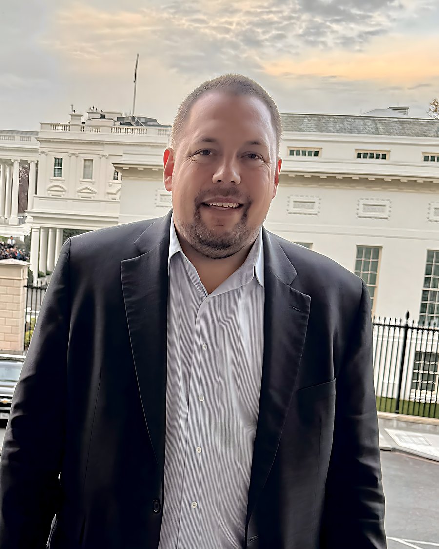 Gregg Roman in a dark blazer on a balcony overlooking the White House West Wing at dusk