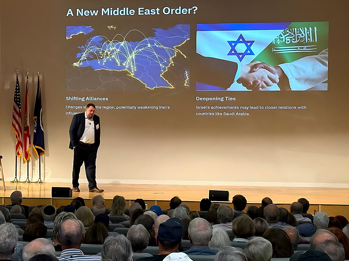 Gregg Roman alone on an auditorium stage beneath a projected slide reading 'A New Middle East Order?'