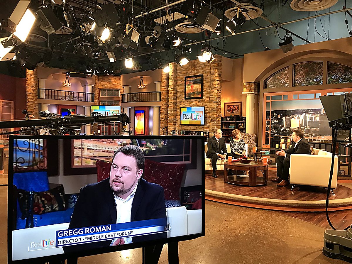 A television studio floor: a foreground monitor shows Gregg Roman in close-up with a 'Gregg Roman — Director, Middle East Forum' chyron while he is interviewed on the set behind