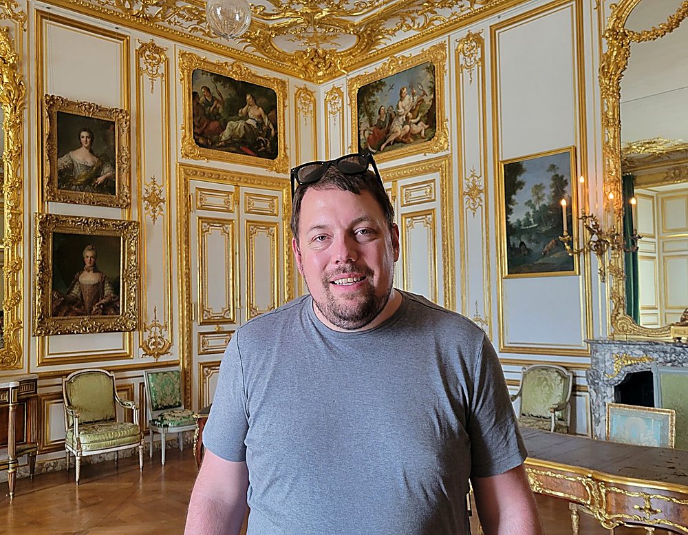 Gregg Roman in a t-shirt with sunglasses on his head, standing in a gilded palace room at Versailles